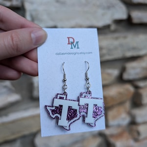 May include: Purple glitter Texas-shaped earrings with a white 'T' on each earring. The earrings are hanging from silver hooks.