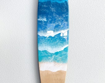 May include: A wooden surfboard with a blue and white resin ocean wave design. The surfboard is hanging on a white wall.