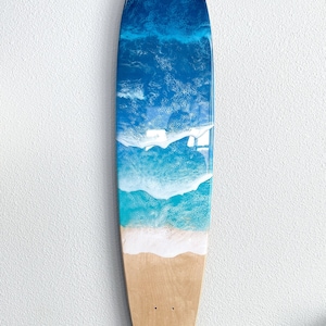 May include: A wooden surfboard with a blue and white resin ocean wave design. The surfboard is hanging on a white wall.