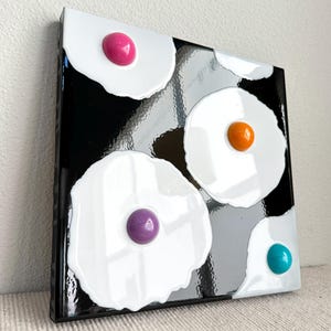 May include: Abstract art piece featuring white, egg-shaped designs with colorful centers against a black and gray background. The centers are various colors, including pink, orange, purple, and teal. The artwork is mounted on a black frame.