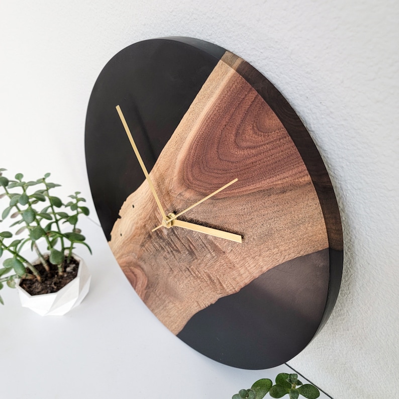May include: A round, black and brown wall clock with gold hands. The clock face is half black and half brown wood with a natural wood grain pattern.