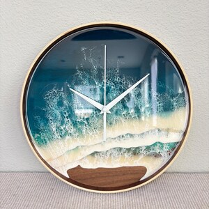 May include: A round wall clock with a wooden frame featuring a resin art design of ocean waves crashing onto a sandy shore. The clock face displays deep blue and turquoise water with white foam details and a dark brown wooden base.