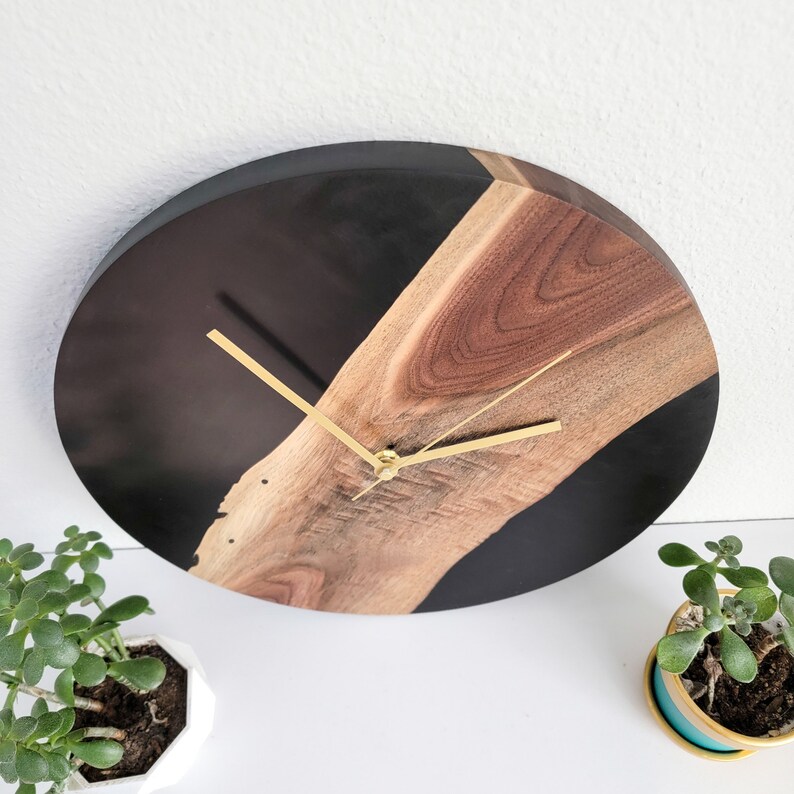 May include: A round, black and brown wall clock with gold hands. The clock face is made of a dark wood with a black epoxy resin filling.