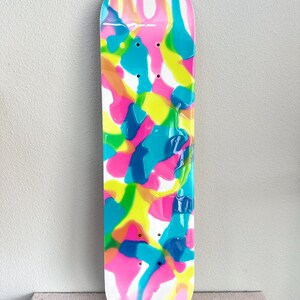 May include: A skateboard with a colorful abstract design. The deck features a white base with vibrant splashes of pink, yellow, blue, and teal. The design is glossy and covers the entire surface of the board.