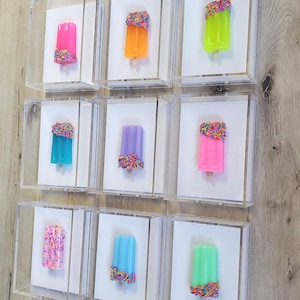 May include: Nine individual clear acrylic shadow boxes, each containing a single popsicle sculpture. The popsicles feature vibrant colors including neon pink, orange, lime green, teal, purple, and light blue, with some decorated with colorful sprinkles.
