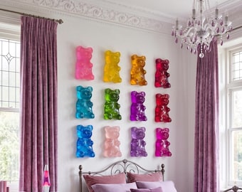 Puede incluir: Una colección de doce esculturas de ositos de goma montadas en la pared y dispuestas en cuadrícula. Cada oso está hecho de un material translúcido y brillante de un color vibrante distinto, incluyendo rosa, amarillo, naranja, rojo, azul, verde y morado.