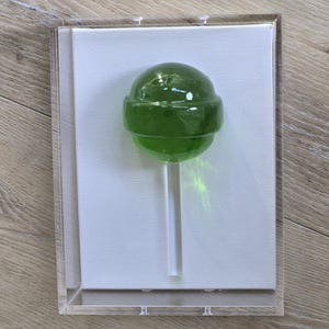 May include: A green lollipop on a clear stick, encased in a clear acrylic box. The lollipop is a vibrant green color and appears to be made of hard candy. The background is a white canvas. The box is displayed on a wooden surface.