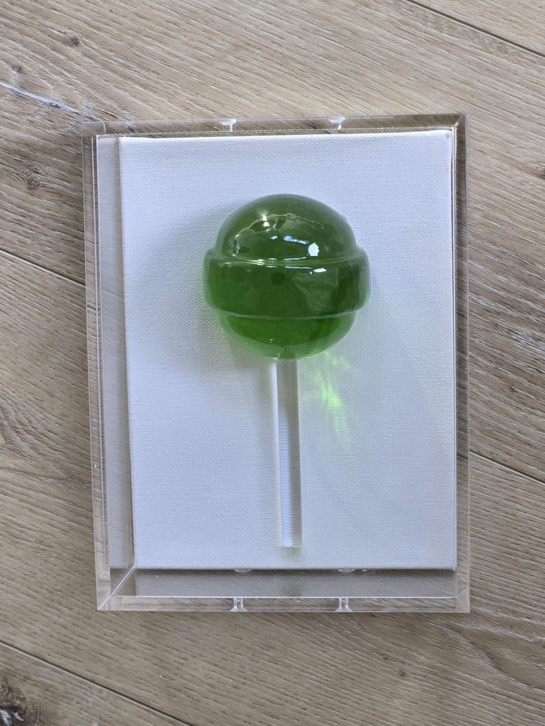 May include: A green lollipop on a clear stick, encased in a clear acrylic box. The lollipop is a vibrant green color and appears to be made of hard candy. The background is a white canvas. The box is displayed on a wooden surface.