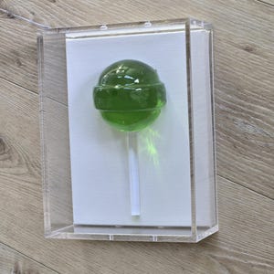 May include: A green lollipop on a white stick, encased in a clear acrylic box. The lollipop is a vibrant green color and appears to be made of hard candy. The box is rectangular and sits on a light-colored wooden surface.