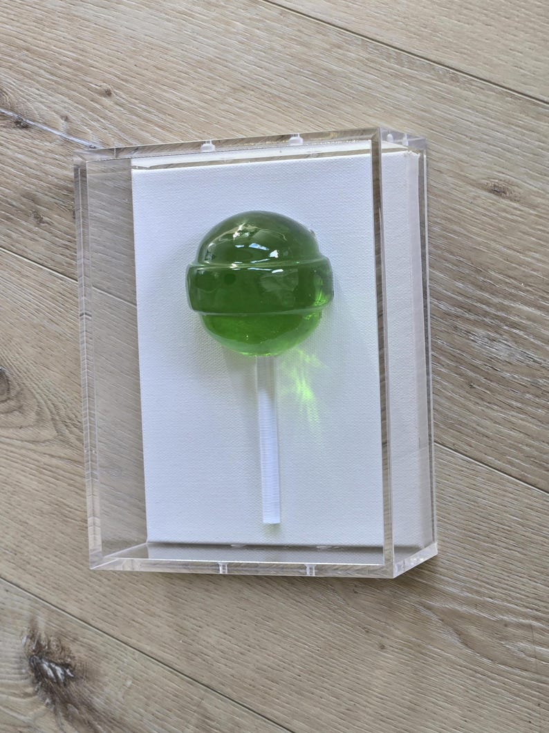 May include: A green lollipop on a white stick, encased in a clear acrylic box. The lollipop is a vibrant green color and appears to be made of hard candy. The box is rectangular and sits on a light-colored wooden surface.