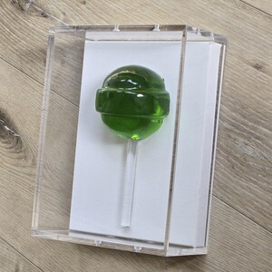 May include: A green lollipop with a clear stick is displayed inside a clear acrylic box. The lollipop is round with a band around the middle. The box is on a light brown wooden surface.