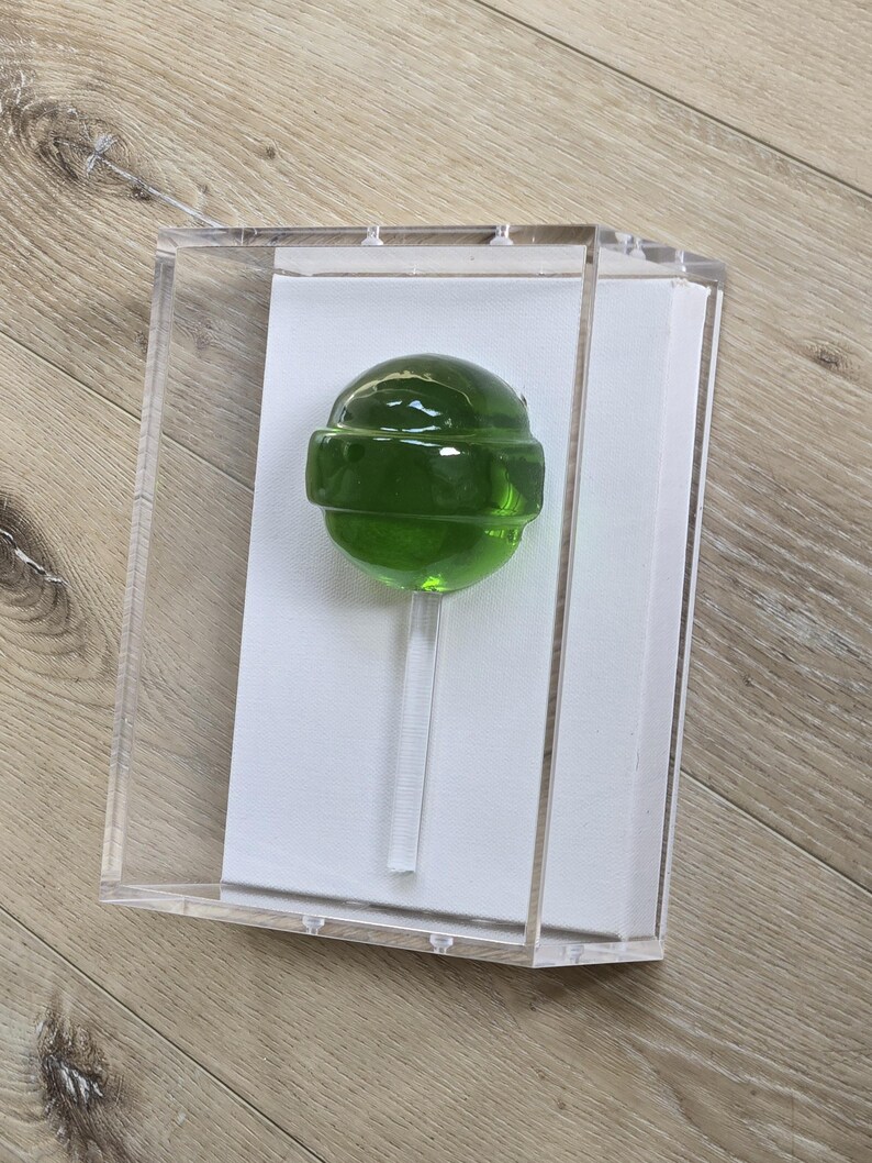 May include: A green lollipop with a clear stick is displayed inside a clear acrylic box. The lollipop is round with a band around the middle. The box is on a light brown wooden surface.