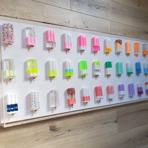 May include: A white wall display featuring a range of colourful popsicle-shaped acrylic sculptures. The popsicles are arranged in rows and feature different colours, patterns, and textures.