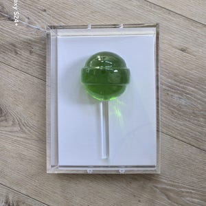 May include: A green lollipop on a white stick, encased in a clear acrylic box. The lollipop has a round shape and a transparent, glossy appearance. The box is displayed on a light wood surface.