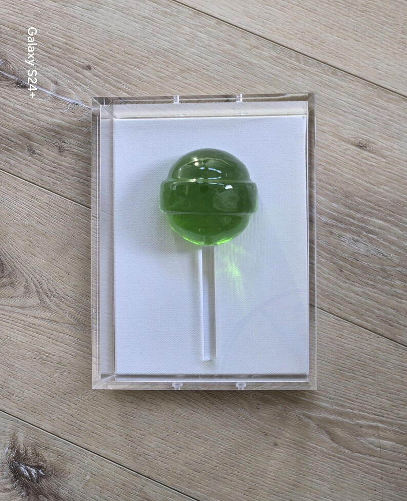May include: A green lollipop on a white stick, encased in a clear acrylic box. The lollipop has a round shape and a transparent, glossy appearance. The box is displayed on a light wood surface.