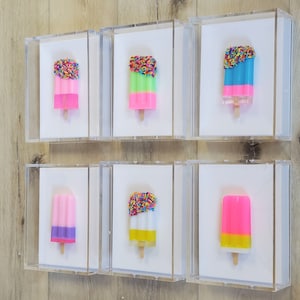 May include: Six framed art pieces featuring colorful popsicle sculptures. Each popsicle has a different color scheme, including pink, green, blue, and yellow, with some topped with rainbow sprinkles. The popsicles are mounted on white backgrounds within clear acrylic boxes.