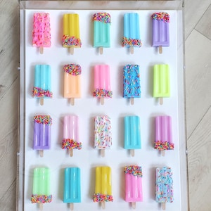 Puede incluir: Una presentación enmarcada de 18 paletas de hielo coloridas con chispas. Las paletas de hielo están dispuestas en tres filas de seis, con cada fila presentando una combinación de colores diferente. Las paletas de hielo están todas hechas de un material liso y brillante y tienen una apariencia realista.