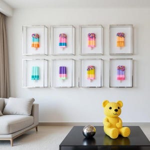 Puede incluir: Ocho obras de arte enmarcadas de paletas heladas con diseños coloridos y chispas. Las paletas se exhiben en cajas transparentes con marcos plateados, montadas en una pared blanca. Un oso de peluche amarillo y una esfera plateada están sobre una mesa negra.