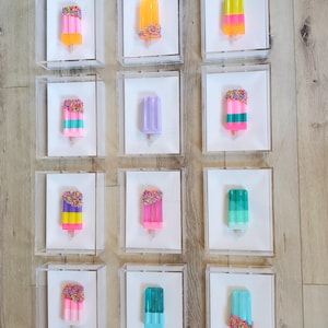 May include: A collection of colorful popsicle art pieces displayed in clear acrylic boxes. Each popsicle features vibrant colors like pink, yellow, green, and blue, some with sprinkles. The artwork is mounted on white backgrounds.