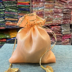 May include: A peach-colored drawstring pouch with gold accents. The pouch is made of a sheer fabric and has gold tassels on the ends of the drawstring. The pouch is sitting on a textured surface. The background is filled with colorful fabrics.