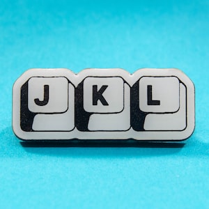 Puede incluir: Un pin de esmalte blanco y negro con tres teclas blancas con las letras J, K y L en negro.