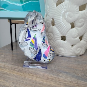 May include: A white oyster shell with a painted design of colorful sailboats. The shell is on a clear acrylic stand.