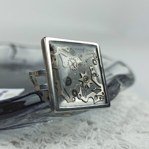 May include: A silver ring with a square clear resin inlay featuring a variety of silver and gold watch gears and cogs.