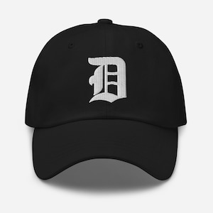 Puede incluir: Gorra de béisbol negra con la letra "D" blanca bordada en una fuente estilizada. La gorra tiene una visera curva y una correa ajustable en la parte trasera. El sombrero está hecho de una tela suave y duradera.