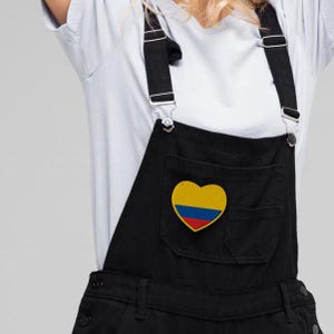 May include: Black denim overalls with adjustable straps and a front pocket. The pocket features a heart-shaped patch with the colors of the Colombian flag: yellow, blue, and red. The overalls are worn over a white t-shirt.