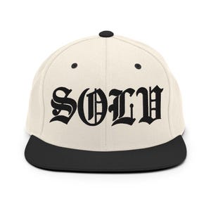 Puede incluir: Una gorra de béisbol color crema con visera plana negra y botón superior negro. El panel frontal presenta la palabra SOLV bordada en una fuente gótica negra y llamativa. La gorra tiene seis paneles con ojales negros para ventilación.