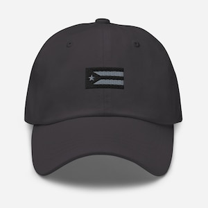 May include: A dark gray baseball cap featuring an embroidered Puerto Rican flag on the front. The flag design includes a white star and horizontal stripes in white and light blue. The cap has a curved brim and a structured crown.
