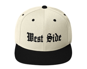 Op de afbeelding: Een beige en zwarte baseballpet met de woorden "West Side" in zwart geborduurd op de voorkant.