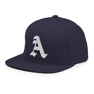 Pode incluir: Boné de beisebol azul marinho com aba reta. O boné apresenta uma letra "A" branca estilizada bordada na frente. O chapéu tem um ajuste estruturado e um fecho de pressão.