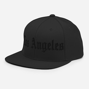 Peut inclure: Casquette de baseball noire avec une visière plate. Le texte "Los Angeles" est brodé en police gothique sur le devant. La casquette a une coupe structurée et un bouton sur le dessus.
