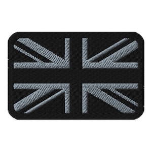 Op de afbeelding: Een rechthoekige patch met het ontwerp van de Union Jack-vlag. Het ontwerp van de vlag, met zijn lichtgrijze kruis en diagonale strepen, is geplaatst tegen een zwarte achtergrond. De patch heeft afgeronde hoeken en een getextureerde afwerking, mogelijk geborduurd of geweven.