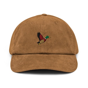 Puede incluir: Gorra de béisbol de pana marrón con visera curva. La gorra presenta una imagen bordada de un pato volador en tonos verdes, rojos, amarillos y negros. Un accesorio de estilo clásico.