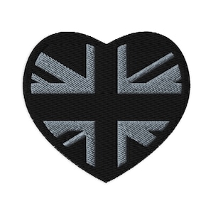 Puede incluir: Un parche bordado en forma de corazón negro, que muestra un diseño de la bandera británica gris. El parche tiene un aspecto texturizado, con los detalles de la bandera cosidos sobre la tela negra. El diseño es una representación estilizada de la bandera británica.