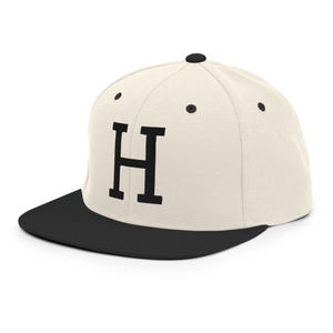 May include: Cream-colored baseball cap with a black brim and the letter "H" in bold black font. The cap has a classic flat brim design and a button on top. A stylish accessory for casual wear.