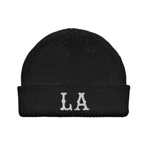 Op de afbeelding: Zwarte gebreide beanie met de witte letters "LA" geborduurd op de voorkant. De beanie heeft een geribbelde manchet en een afgeronde bovenkant. De letters zijn in een gestileerd lettertype.