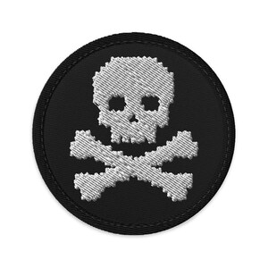Könnte beinhalten: Runder, gestickter Aufnäher mit einem weißen Totenkopf und gekreuzten Knochen auf schwarzem Hintergrund. Der Aufnäher hat einen gestickten Rand. Der Totenkopf und die gekreuzten Knochen sind detailliert.