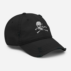 Peut inclure: Casquette de baseball noire et vieillie, ornée d'un motif de tête de mort et d'os croisés blancs. La casquette a une visière incurvée et un aspect usé, avec des bords effilochés. Le motif est brodé sur le devant.