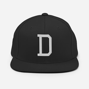 Puede incluir: Gorra de béisbol negra con una visera plana y una letra "D" blanca bordada en la parte delantera. La gorra tiene un ajuste estructurado y un botón en la parte superior.