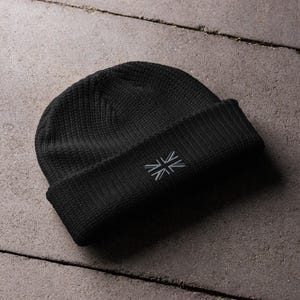 May include: A black knit beanie with a folded brim. The beanie features a small, embroidered Union Jack flag on the front. The hat is made of a textured material and is suitable for cold weather.