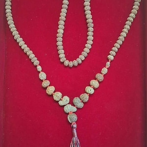 Könnte beinhalten: Eine Perlenkette mit Quaste, präsentiert auf roter Oberfläche. Die Kette besteht aus zahlreichen kleinen, runden Perlen in verschiedenen Grau- und Brauntönen. Die Quaste besteht aus roten, blauen und schwarzen Fäden.