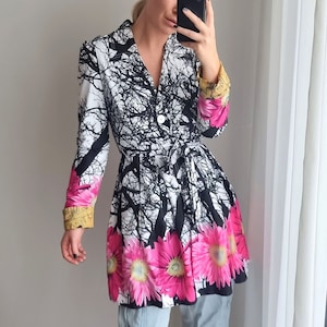 May include: A white and black floral print jacket with a pink and yellow floral trim. The jacket has a cinched waist and a long, flowing skirt. The model is wearing light blue jeans and black heeled shoes.