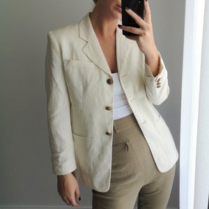 May include: A white linen blazer with a button closure and two pockets. The blazer is worn over a white tank top and brown pants.