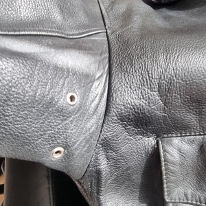 Op de afbeelding: Close-up van een zwart leren jack met een rits en twee zilveren drukknopen op de mouw.