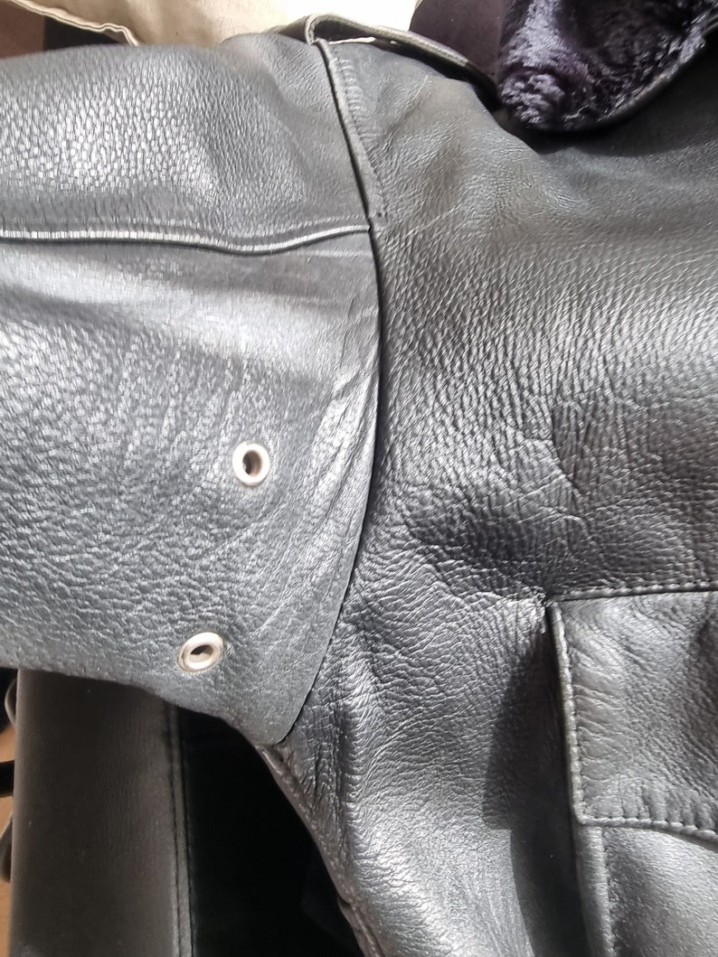 Op de afbeelding: Close-up van een zwart leren jack met een rits en twee zilveren drukknopen op de mouw.