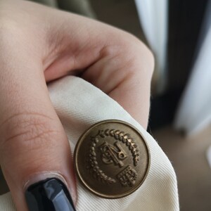 May include: A close-up of a gold-colored button with a laurel wreath design and the letters "BP" in the center.