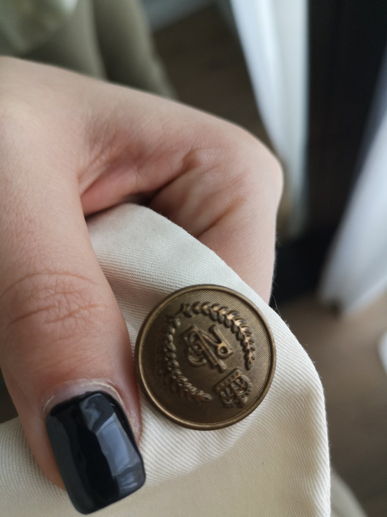 May include: A close-up of a gold-colored button with a laurel wreath design and the letters "BP" in the center.